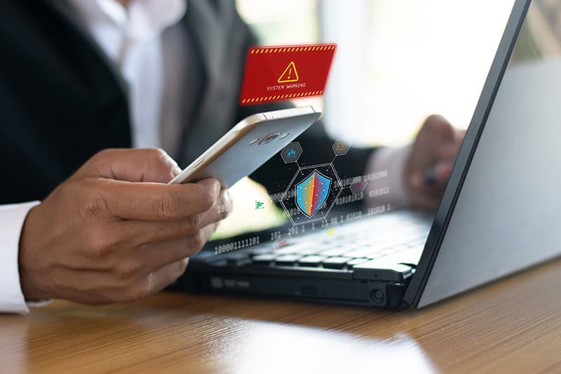 Person holding a smartphone with a security alert on screen, connected to a laptop.
